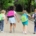 Four young children walk hand in hand down a tree-lined path, each wearing a backpack. The second child from the right has a backpack with the text "BACK TO SCHOOL" written on it. The kids, seen from behind, appear happy and ready for school, with the surrounding greenery suggesting a late summer or early fall day.