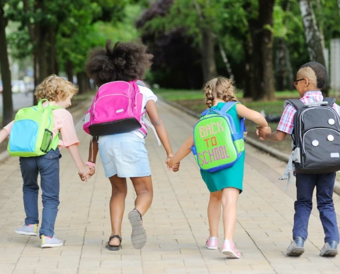 Four young children walk hand in hand down a tree-lined path, each wearing a backpack. The second child from the right has a backpack with the text "BACK TO SCHOOL" written on it. The kids, seen from behind, appear happy and ready for school, with the surrounding greenery suggesting a late summer or early fall day.