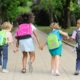 Four young children walk hand in hand down a tree-lined path, each wearing a backpack. The second child from the right has a backpack with the text "BACK TO SCHOOL" written on it. The kids, seen from behind, appear happy and ready for school, with the surrounding greenery suggesting a late summer or early fall day.