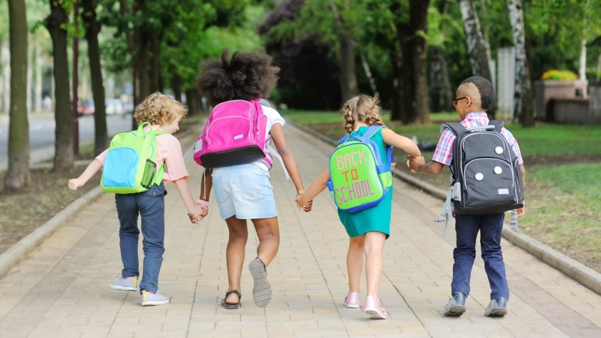 Four young children walk hand in hand down a tree-lined path, each wearing a backpack. The second child from the right has a backpack with the text "BACK TO SCHOOL" written on it. The kids, seen from behind, appear happy and ready for school, with the surrounding greenery suggesting a late summer or early fall day.