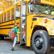 A young child with short red hair and a large camouflage-patterned backpack steps onto a yellow school bus. The bus door is open, and the child is lifting a foot onto the first step. The bus is parked on a tree-lined suburban street, with the sun casting a warm glow through the trees. Other students can be seen sitting inside the bus through the windows.