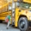 A young child with short red hair and a large camouflage-patterned backpack steps onto a yellow school bus. The bus door is open, and the child is lifting a foot onto the first step. The bus is parked on a tree-lined suburban street, with the sun casting a warm glow through the trees. Other students can be seen sitting inside the bus through the windows.