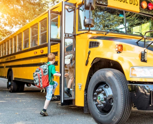 A young child with short red hair and a large camouflage-patterned backpack steps onto a yellow school bus. The bus door is open, and the child is lifting a foot onto the first step. The bus is parked on a tree-lined suburban street, with the sun casting a warm glow through the trees. Other students can be seen sitting inside the bus through the windows.