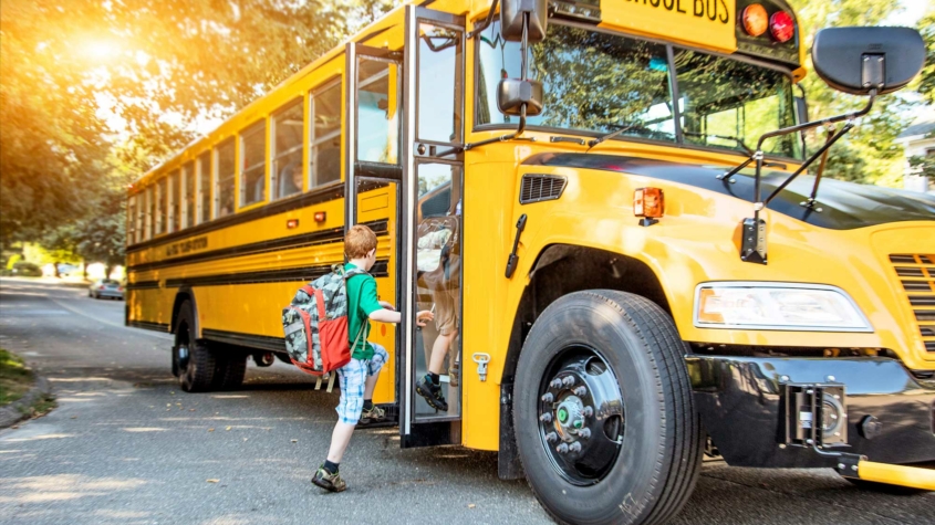 A young child with short red hair and a large camouflage-patterned backpack steps onto a yellow school bus. The bus door is open, and the child is lifting a foot onto the first step. The bus is parked on a tree-lined suburban street, with the sun casting a warm glow through the trees. Other students can be seen sitting inside the bus through the windows.