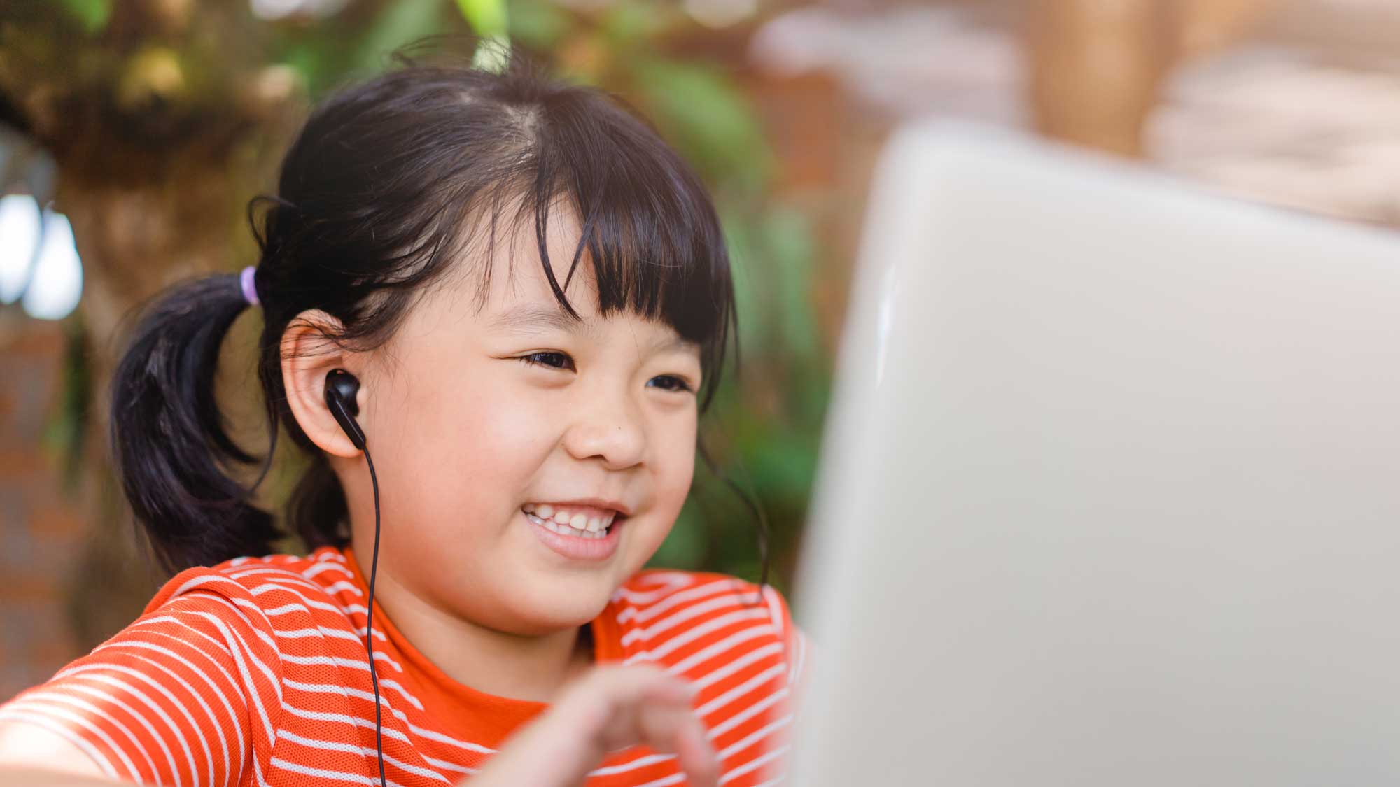 A young girl with black hair in pigtails is smiling while looking at a laptop screen. She is wearing a red and white striped shirt and has black earbuds in her ears. The background is softly blurred with greenery, suggesting she is outdoors.