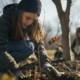 A young woman wearing a beanie and gloves crouches down to plant a small tree sapling in the soil. The sapling has a single dried leaf still attached. She is focused on her task, surrounded by other volunteers who are also planting trees in a park setting. The background shows bare trees and soft sunlight, suggesting an autumn or early winter day.