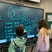 Two children stand at a large interactive screen in a classroom, writing words that follow the "a_e" vowel-consonant-e pattern (e.g., "late," "mate," "plate"). Both kids use styluses, one writing on the left side and the other on the right. The classroom has educational supplies, including books, trays, and a cart labeled "Ms. Grap." A sign in the background reads, "You need to calm down, you're being too loud."