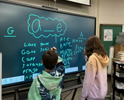 Two children stand at a large interactive screen in a classroom, writing words that follow the "a_e" vowel-consonant-e pattern (e.g., "late," "mate," "plate"). Both kids use styluses, one writing on the left side and the other on the right. The classroom has educational supplies, including books, trays, and a cart labeled "Ms. Grap." A sign in the background reads, "You need to calm down, you're being too loud."