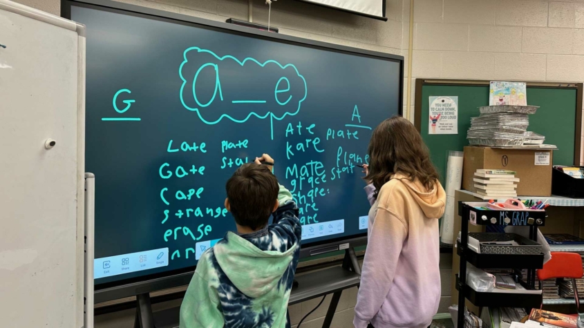 Two children stand at a large interactive screen in a classroom, writing words that follow the "a_e" vowel-consonant-e pattern (e.g., "late," "mate," "plate"). Both kids use styluses, one writing on the left side and the other on the right. The classroom has educational supplies, including books, trays, and a cart labeled "Ms. Grap." A sign in the background reads, "You need to calm down, you're being too loud."