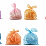 An arrangement of eight plastic garbage bags in various colors is shown. The top row includes purple, light pink, orange, and red bags. The bottom row features light blue, orange, light blue again, and lavender bags. Each bag is tied at the top, with slight variations in size and fullness. The bags are displayed against a white background.