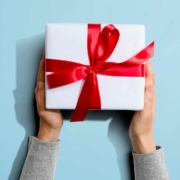 A pair of hands holds a neatly wrapped white gift box with a shiny red ribbon and bow. The background is a light blue, providing contrast to the bright red ribbon. The hands are positioned symmetrically, and the gift appears centered in the frame. The person's sleeves are visible, suggesting they are wearing a light gray shirt.