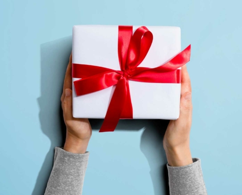 A pair of hands holds a neatly wrapped white gift box with a shiny red ribbon and bow. The background is a light blue, providing contrast to the bright red ribbon. The hands are positioned symmetrically, and the gift appears centered in the frame. The person's sleeves are visible, suggesting they are wearing a light gray shirt.