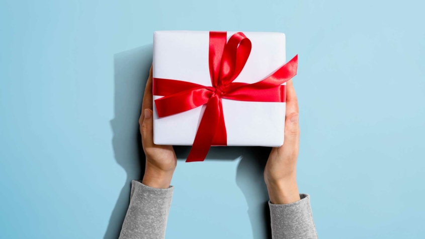 A pair of hands holds a neatly wrapped white gift box with a shiny red ribbon and bow. The background is a light blue, providing contrast to the bright red ribbon. The hands are positioned symmetrically, and the gift appears centered in the frame. The person's sleeves are visible, suggesting they are wearing a light gray shirt.