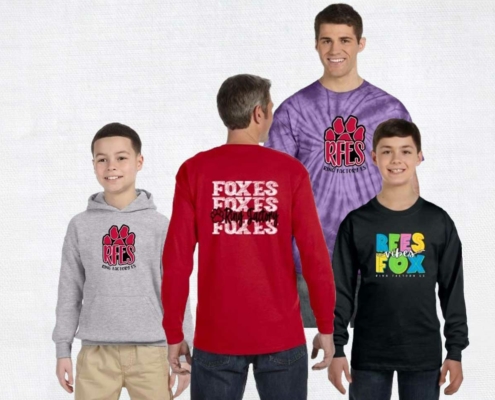 Four individuals modeling Ring Factory Elementary spiritwear. From left to right: a child wearing a gray hoodie with a red paw print and "RFES," a person in a red long-sleeve shirt with the word "Foxes" repeated on the back, a man in a purple tie-dye shirt with the same paw print and "RFES" logo, and another child in a black long-sleeve shirt with colorful text that reads "RFES Vibes Fox."