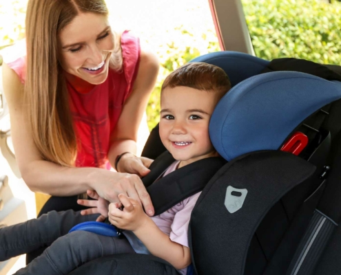 A smiling woman helps secure a young child in a car seat inside a vehicle. The child, also smiling, is seated in a black and blue car seat and is being buckled in by the woman. Sunlight enters through the car door, and greenery is visible outside the window.