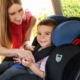 A smiling woman helps secure a young child in a car seat inside a vehicle. The child, also smiling, is seated in a black and blue car seat and is being buckled in by the woman. Sunlight enters through the car door, and greenery is visible outside the window.