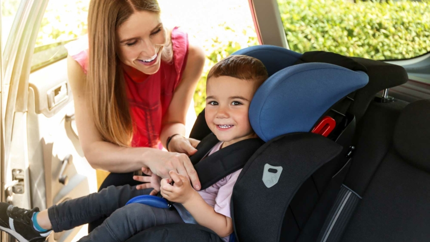 A smiling woman helps secure a young child in a car seat inside a vehicle. The child, also smiling, is seated in a black and blue car seat and is being buckled in by the woman. Sunlight enters through the car door, and greenery is visible outside the window.