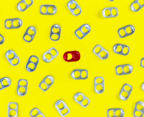 A pattern of silver soda can tabs scattered across a bright yellow background, with one distinctive red tab placed near the center, standing out among the silver tabs.