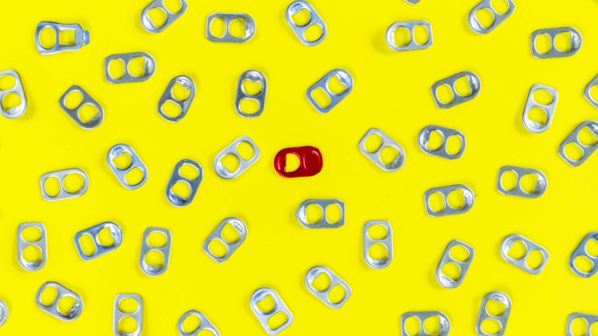 A pattern of silver soda can tabs scattered across a bright yellow background, with one distinctive red tab placed near the center, standing out among the silver tabs.