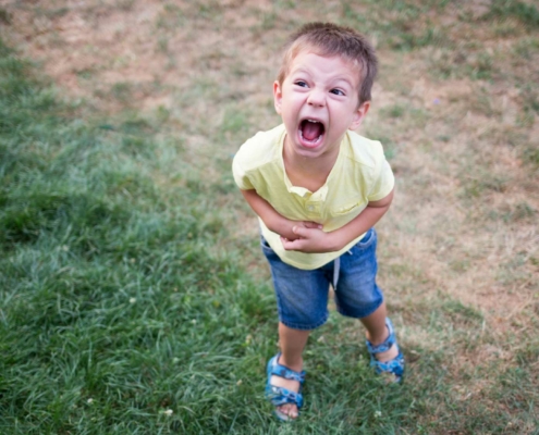 A young boy in a yellow shirt and blue shorts stands on a patchy grassy lawn, shouting with an exaggerated facial expression. He holds his stomach as if pretending to be in distress or acting playful. His sandals are blue, and his posture is slightly leaned forward, emphasizing his animated demeanor.