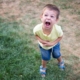 A young boy in a yellow shirt and blue shorts stands on a patchy grassy lawn, shouting with an exaggerated facial expression. He holds his stomach as if pretending to be in distress or acting playful. His sandals are blue, and his posture is slightly leaned forward, emphasizing his animated demeanor.