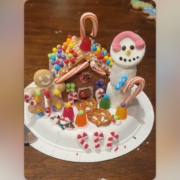 A festive gingerbread house on a paper plate decorated with colorful candies, pretzels, and candy canes. To the left is a gingerbread figure holding a candy cane, and to the right is a marshmallow snowman with a pink icing scarf, a candy cane arm, and a smiling face made of icing. Gumdrops, sprinkles, and small peppermint decorations surround the scene on a wooden table.