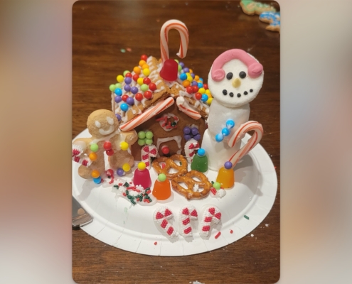 A festive gingerbread house on a paper plate decorated with colorful candies, pretzels, and candy canes. To the left is a gingerbread figure holding a candy cane, and to the right is a marshmallow snowman with a pink icing scarf, a candy cane arm, and a smiling face made of icing. Gumdrops, sprinkles, and small peppermint decorations surround the scene on a wooden table.