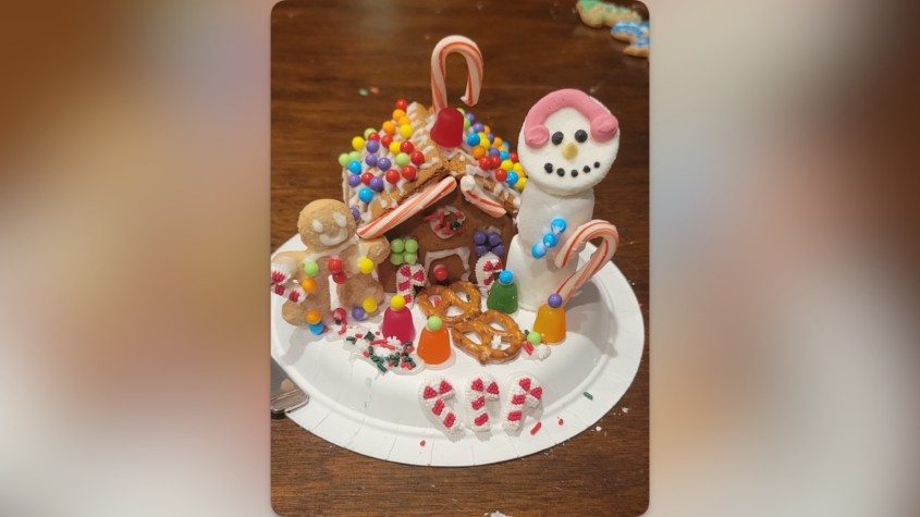 A festive gingerbread house on a paper plate decorated with colorful candies, pretzels, and candy canes. To the left is a gingerbread figure holding a candy cane, and to the right is a marshmallow snowman with a pink icing scarf, a candy cane arm, and a smiling face made of icing. Gumdrops, sprinkles, and small peppermint decorations surround the scene on a wooden table.